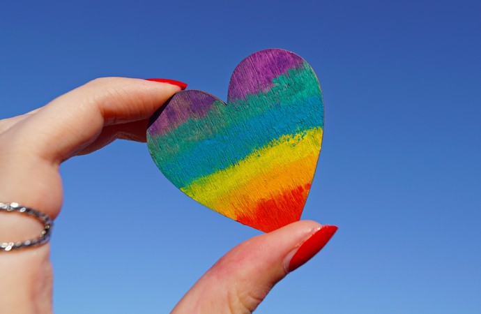 ein Herz in Regenbogenfarben wird von zwei Fingern in den Himmel gehalten